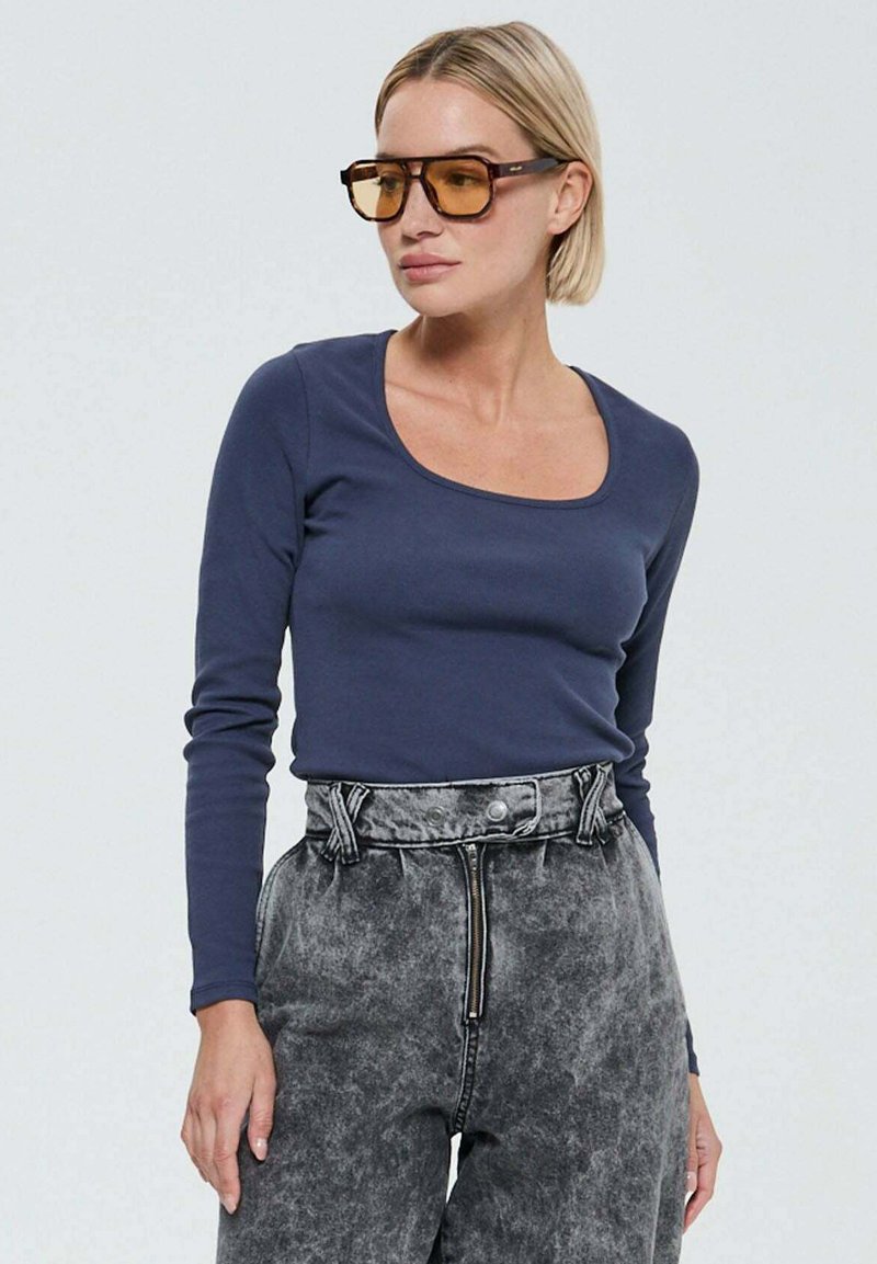 Haut à manches longues bleu marine avec un col en scoop, associé à un pantalon gris à taille haute en délavé acide ; le mannequin porte de grandes lunettes teintées.