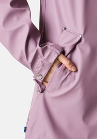 Giacca impermeabile di un delicato viola, caratterizzata da un polsino aderente con bottoni a pressione e una tasca laterale, realizzata in materiale liscio e resistente.