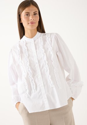 Jonge vrouw met steil bruin haar, een witte blouse met ruches en beige broek, handen in de zakken, neutrale achtergrond.