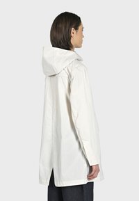 Giacca leggera bianca con cappuccio, maniche lunghe, apertura posteriore e silhouette dritta. Presenta un design semplice e una texture liscia.