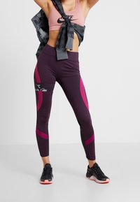 Leggings sportivi viola con dettagli rosa e neri, caratterizzati da un design aderente e una texture liscia. Abbinati a un reggiseno sportivo rosa e sneakers nere.