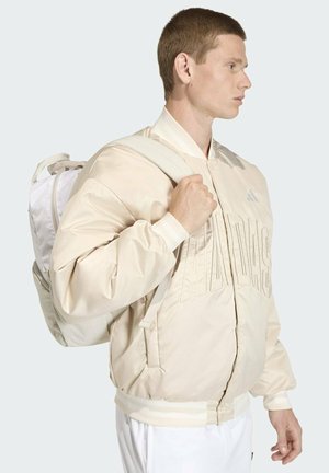 Beige bomber jacket with ribbed cuffs and collar, featuring embossed logo. The model carries a light-colored backpack with padded straps.