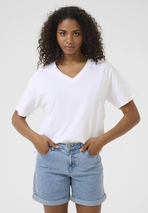 Femme aux cheveux bouclés portant un t-shirt blanc à col en V et un short en jean bleu clair retroussé, mains dans les poches.