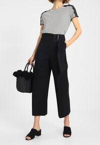 Černé široké kalhoty s páskem v pase, doplněné pruhovaným černobílým tričkem s černými krajkovými akcenty; černé skluzné sandály; texturovaná kabelka.