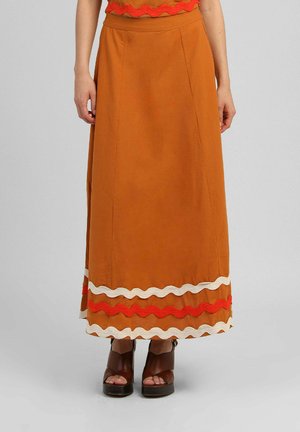 Longue jupe orange brûlé avec un bord ondulé rouge et blanc près de l'ourlet, portée avec des sandales à plateforme marron sur fond gris clair.