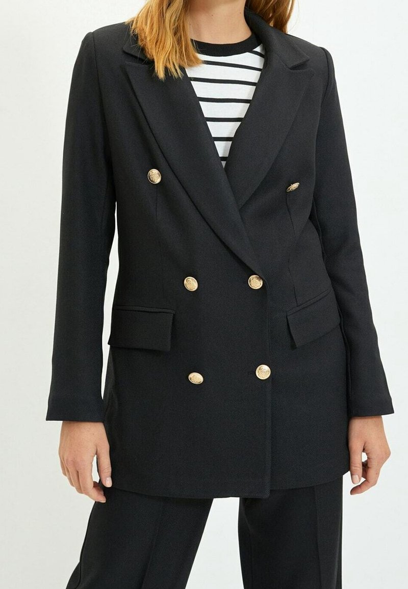 Svart dubbelknäppt blazer med guldfärgad knappdetaljer, två framfickor, slagkrage och textil med struktur. Randig skjorta under.