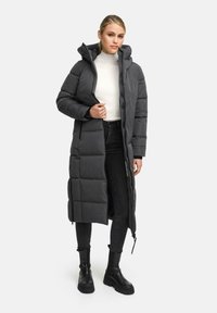 Giacca a vento lunga grigio scuro con cappuccio, caratterizzata da una chiusura con zip e un design a blocchi trapuntato, abbinata a jeans skinny neri e stivali robusti.