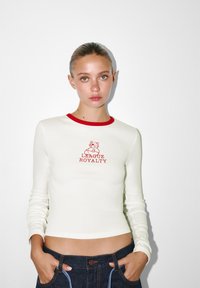 Pitkähihaisen valkoisen paidan punaisella kauluksella, jonka graafisessa printissä on karhu ja teksti "LEAGUE ROYALTY" punaisella. Sileä pinta, istuva malli.