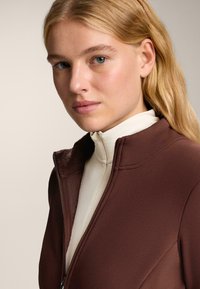 Smeđi zip-jakna s visokim ovratnikom, s mekanim unutarnjim podstavom. Vanjski materijal je gladak, dizajniran za udobnost i toplinu.