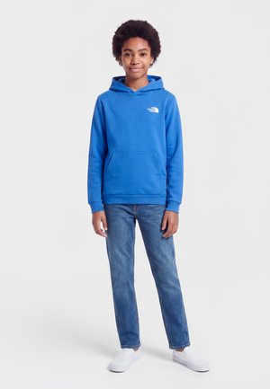 Jeune personne debout, face à nous, portant un sweat à capuche bleu, un jean bleu et des chaussures blanches sans lacets sur un fond clair uni.