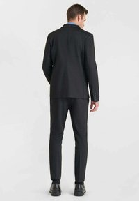 Completo nero su misura in tessuto strutturato, con giacca a risvolto e pantaloni slim-fit. Completato con dettagli in bottone e scarpe eleganti.