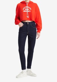 Sudadera corta roja con texto blanco, combinada con jeans ajustados de talle alto en azul oscuro. El modelo lleva zapatillas blancas con cordones visibles.