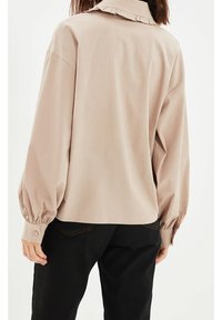 Femme portant une blouse beige à manches longues avec un détail de col plissé, associée à un pantalon noir, vue de dos.