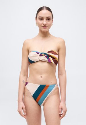 Femeie purtând un bikini bandeau fără bretele, multicolor, cu o cataramă circulară și slipi asortați cu tăietură înaltă, stând pe un fundal simplu deschis la culoare.