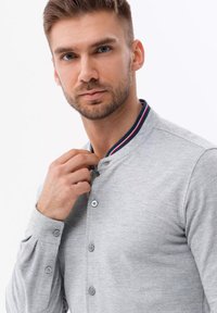 Lichtgrijs shirt met kraag, voorzien van rode en marineblauwe strepen, knoopsluiting aan de voorkant en lange mouwen met knopen bij de manchet. Zachte textuur.