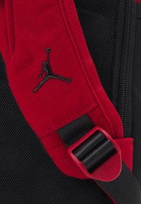 Piros és fekete szövet hátizsák textured felülettel, fekete Jumpman logóval és piros csattal egy fekete hevederen.