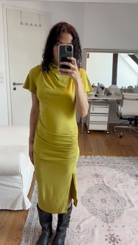 Pessoa vestindo um vestido midi amarelo mostarda com fenda lateral e botas pretas até ao joelho, tirando uma selfie ao espelho em uma sala com piso de madeira.