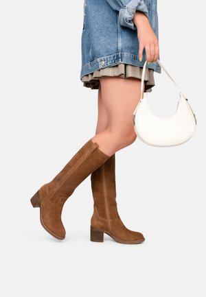 Bottes en daim marron, hautes jusqu'au genou, avec un talon carré, présentant une fermeture éclair sur le côté et une finition lisse. Portées avec une veste en jean et une jupe à carreaux.