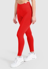 Leggings rossi a vita alta realizzati in tessuto elastico, caratterizzati da una superficie liscia e una silhouette aderente. Indossati con sneaker bianche.
