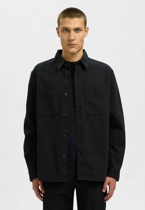 Young man with short hair wearing a black button-up shirt over a black t-shirt, standing against a plain light background.
