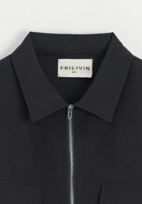 Veste noire avec fermeture éclair à l'avant, col pointu et deux poches poitrine. Fabriquée en tissu lisse ; étiquette de taille visible.