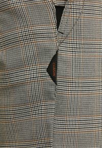 Detailní záběr na šedou a hnědou kostkovanou látku s malou černou štítkou přišitou uvnitř, na které je částečně vidět oranžový text.