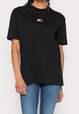 T-shirt nera in cotone con maniche corte, caratterizzata da una piccola toppa logo colorata sul petto, abbinata a jeans in denim blu.