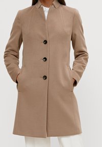 Beige frakke med et struktureret design, som har høj krave, lange ærmer, fire sorte knapper og sidelommer. Glat stoftekstur.