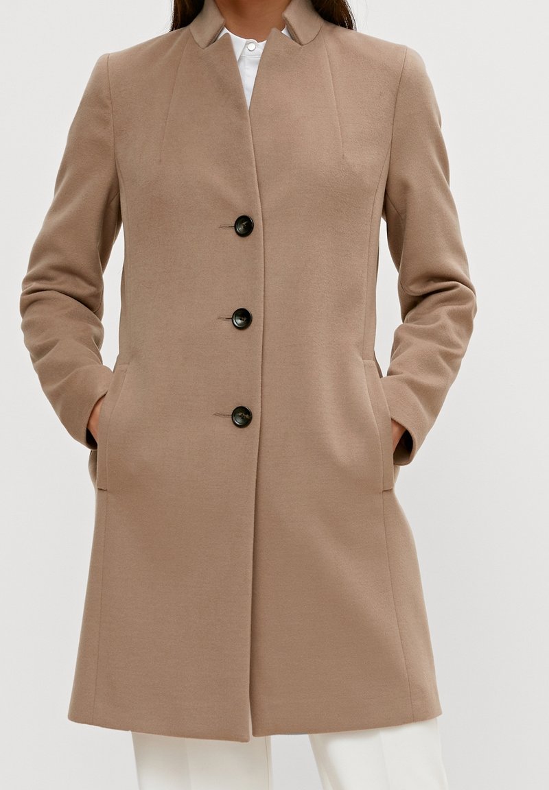 Beige frakke med et struktureret design, som har høj krave, lange ærmer, fire sorte knapper og sidelommer. Glat stoftekstur.