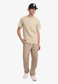 Camiseta de algodón beige combinada con pantalones de tono marrón claro, usada con una gorra negra que presenta un emblema de estrella blanca. Calzado casual completa el conjunto.