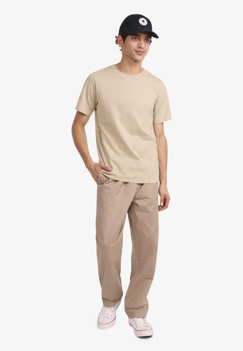 Camiseta de algodón beige combinada con pantalones de tono marrón claro, usada con una gorra negra que presenta un emblema de estrella blanca. Calzado casual completa el conjunto.