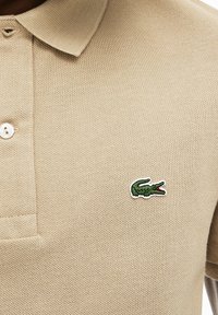Polo de color beige hecho de tela texturizada, con un cierre de tres botones y un pequeño logo de cocodrilo verde en el lado izquierdo del pecho.