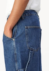 Pantalon en denim avec une coupe décontractée, de couleur bleu profonde, taille élastique, poches latérales et couture contrastante orange sur les coutures.