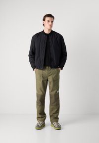 Una chaqueta bomber negra acolchada sobre una camiseta negra, combinada con pantalones cargo verde oliva con parches en las rodillas y zapatillas gris-verdosas.