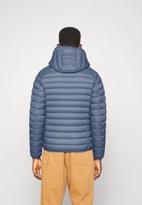 Navy blue puffer jacket with horizontal quilted design, a hood, and elastic cuffs, paired with light brown pants. Smooth texture.