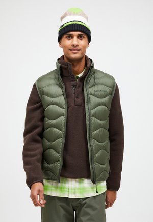 Groene puffer vest met gequiltde textuur, bruine fleece trui eronder, en een gekleurde gebreide beanie. Gelaagde look met een geruite shirt.
