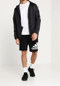 Chaqueta ligera negra con capucha, camiseta negra y pantalones cortos negros con un gran logo blanco de Adidas; combinados con zapatillas negras y calcetines blancos.
