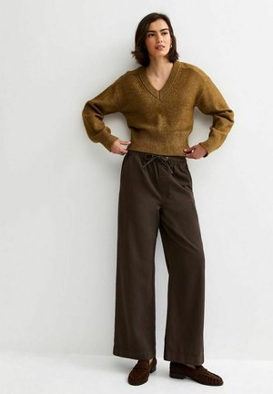 Femme portant un pull marron à col en V, un pantalon large sombre avec taille à cordon, et des mocassins foncés, debout devant un mur blanc uni.