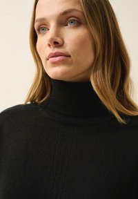 Czarny sweter typu golf z miękką fakturą, ozdobiony subtelnym pionowym przeszyciem. Jasnobrązowe włosy modelki opadają wokół dekoltu.