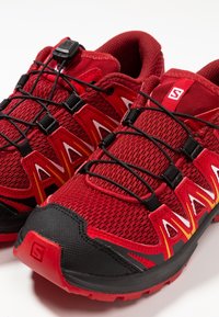 Red athletic shoes with a textured mesh upper, black accents, a secure lacing system, and a durable rubber sole featuring a traction pattern.