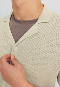 Cardigan beige chiaro con un design a maglia testurizzata, caratterizzato da un colletto e una chiusura con un singolo bottone. Sotto è di colore marrone.