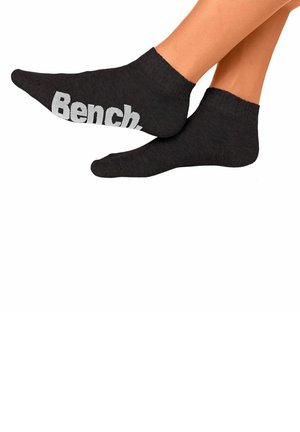 Füße mit schwarzen Knöchelsocken mit „Bench“ in großen grauen Buchstaben auf der Sohle vor weißem Hintergrund.