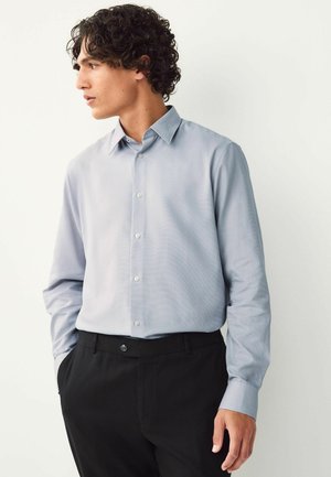 Cămașă albastră deschis cu un model discret în carouri, mâneci lungi și guler ascuțit, purtată cu pantaloni negri.