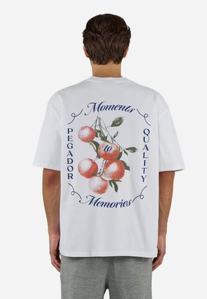 Άνδρας που φοράει λευκό μπλουζάκι με κόκκινο γραφικό κεράσι και κείμενο "Moments to Memories," "Pegador," και "Quality" στην πλάτη.