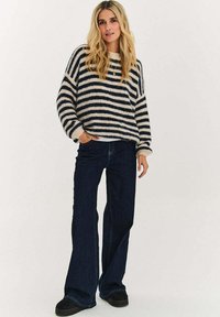 Sweter w paski w kolorze kremowym i granatowym, o luźnym kroju i szerokich rękawach, zestawiony z ciemnoniebieskimi jeansami dzwonami i czarnymi butami.