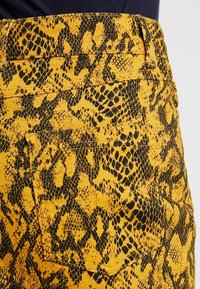 Tela amarilla con un patrón de estampado de serpiente negra, con costuras visibles y un bolsillo trasero. La textura parece suave y ligeramente tejida.