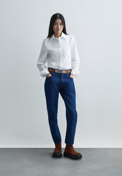 Naine seisab käed taskutes, seljas valge nööbiga särk, sinised teksad Moschino vööga ja pruunid platvormkingad, ühevärvilise tausta ees.