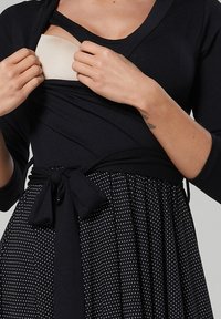 Vestido negro con mangas largas y un cuerpo ajustado, que presenta una falda de lunares y un detalle de cintura atada. Tejido suave con elementos de diseño en capas.