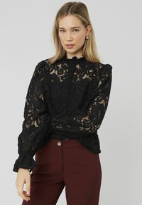 Blouse en dentelle noire avec motifs floraux, manches bouffantes et col haut avec fermeture à boutons, associée à un pantalon bordeaux.