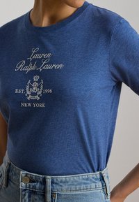 Μπλε μπλουζάκι Lauren Ralph Lauren με λευκό λογότυπο και κείμενο "EST. 1996 NEW YORK", φορεμένο μέσα σε ανοιχτόχρωμο τζιν.